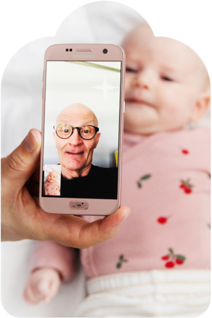 Etualalla älypuhelin, jossa on videopuhelu käynnissä. Puhelussa on kalju silmälasipäinen mies. Puhelimen takana vauva, jolla on yllään vaaleanpunainen paita.